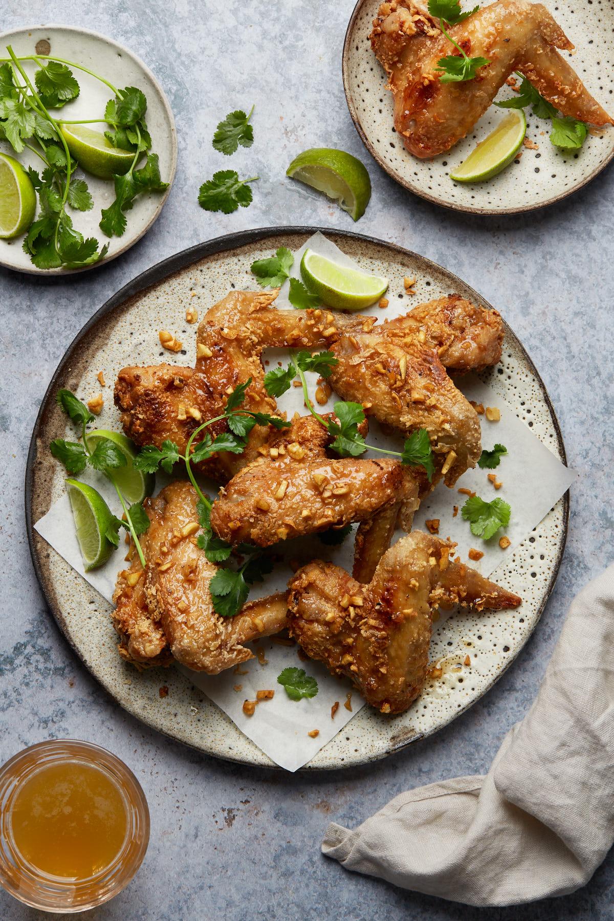 a platter of chicken wings next to a glass of beer, kitchen linen, plate with cilantro and lime wedges, and a plate with one wing